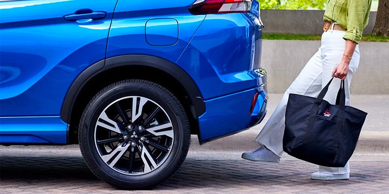 Image of a blue 2024 Mitsubishi Eclipse Cross showcasing the rear driver's side of the vehicle, with a woman's foot underneath the vehicle demonstrating the hands-free power tailgate